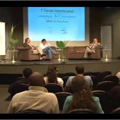 Foto de um salão com pessoas sentadas observando três pessoas em cima de um palco, essas três pessoas estão interagindo entre si e estão sentadas em sofás diferentes.