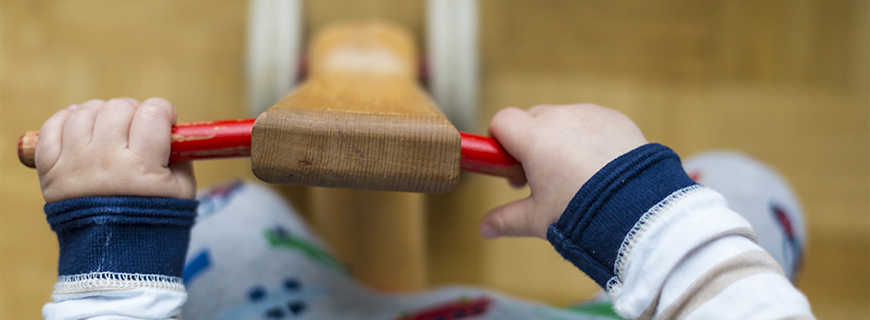 foto em detalhe de mãos de uma criança segurando no guidão de um patinete de madeira