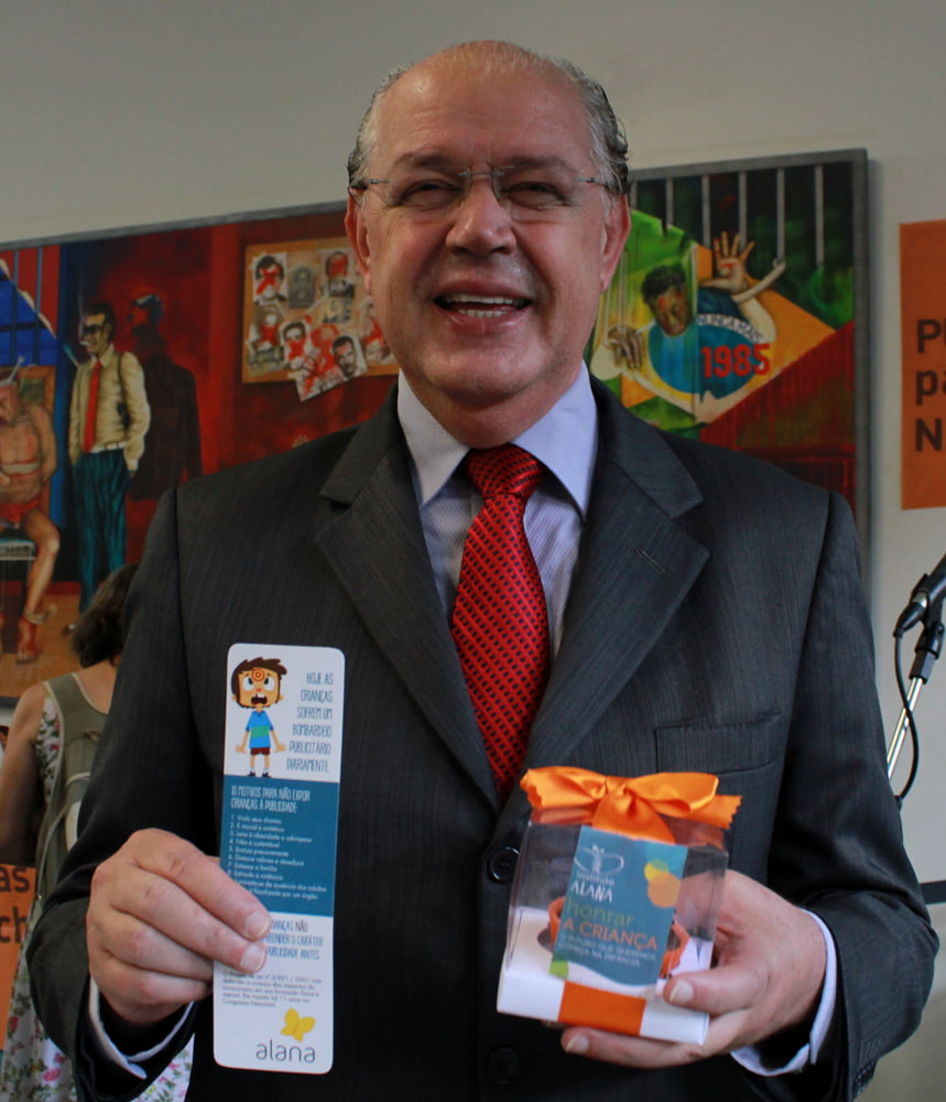 Foto de Luiz Carlos Hauly segurando um panfleto e um presente do Instituto Alana.