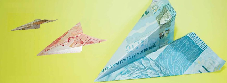 Imagem de aviões de papel feita com notas de dinheiro, uma nota de cem, uma de dez e uma de vinte reais.