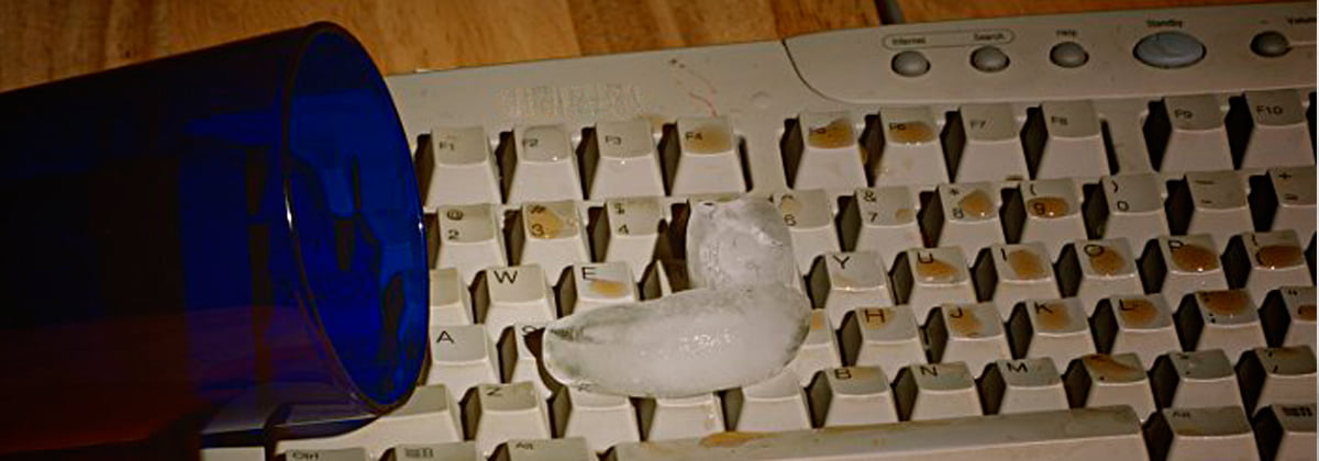 Foto de um copo que derramou bebida em cima de um teclado, ainda tem dois gelos em cima do teclado.