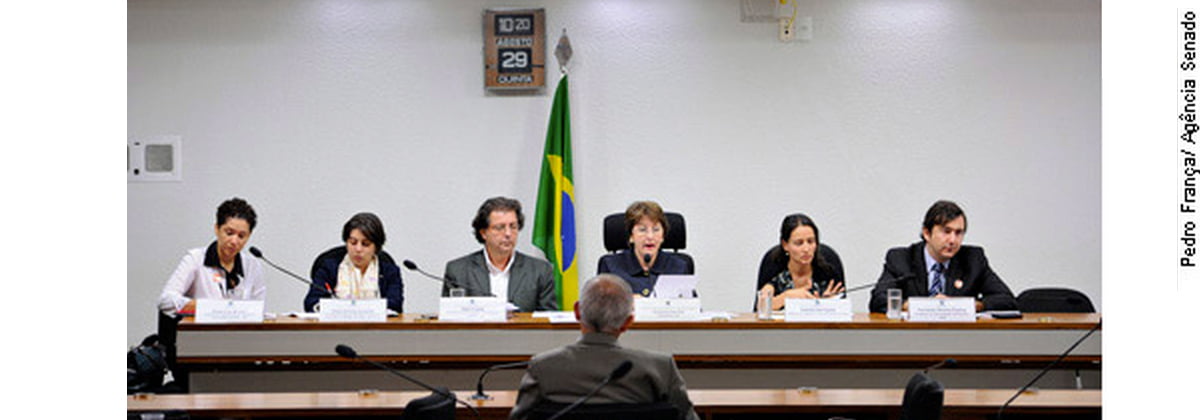 Foto de pessoas sentadas em uma mesa, uma mulher está falando em um microfone nesta mesa, no total há quatro mulheres e dois homens.