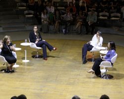 Foto de quatro pessoas, três mulheres e um homem, em um palco sentados em cadeiras, uma das mulheres fala ao microfone.