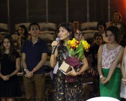 Foto de pessoas em volta de uma mulher falando em um microfone com sua mão direita e segura flores com sua mão esquerda.