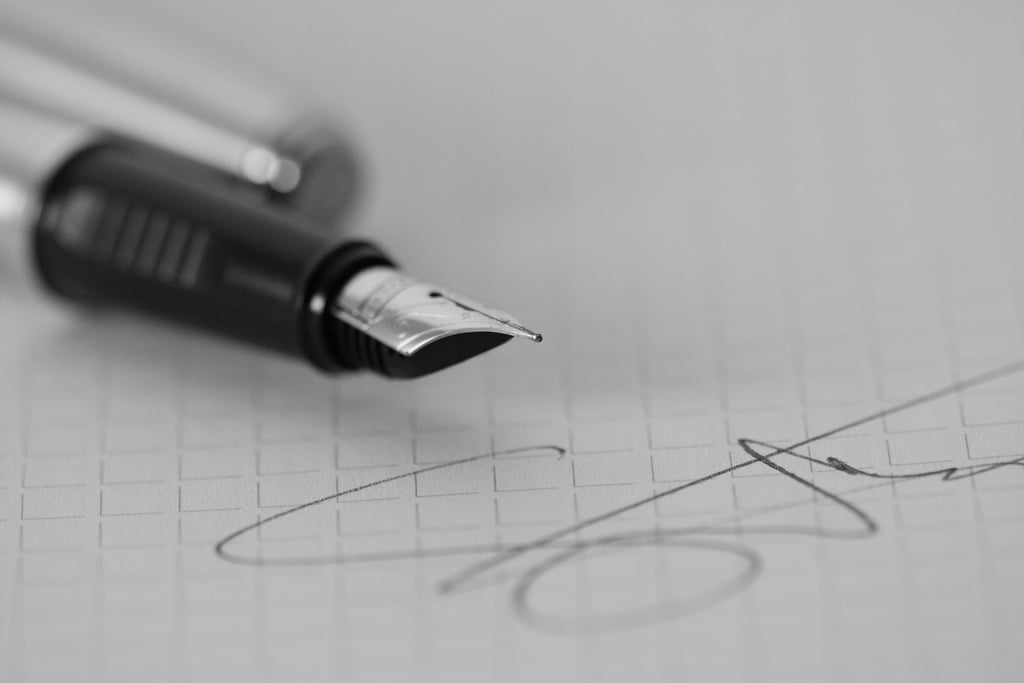 Imagem de uma assinatura em uma folha de papel, tem uma caneta em cima desta folha.