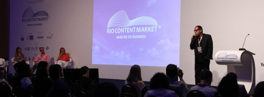 Foto de uma palestra onde pessoas sentadas em um plateia observam um homem em cima de um palco falando a um microfone, um telão no palco descreve: Rio Content Market, Here we do business.
