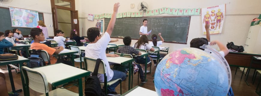 Foto de uma sala de aula um professor está se virando, crianças sentadas em carteiras, alguma levantam o braço.
