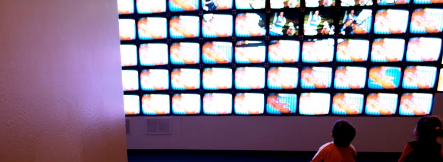 Foto de duas crianças em frente a várias televisões empilhadas, as televisões estão passando algum tipo de programa.