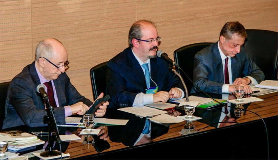 Foto com três homens de terno sentados em uma mesa, um homem fala ao microfone enquanto outros dois estão confirmando documento a mesa.