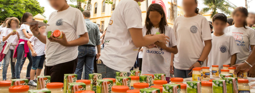 Foto de crianças que formam um fila indiana para receber um suco e um pote de maionese, uma pessoa está distribuindo os produtos.