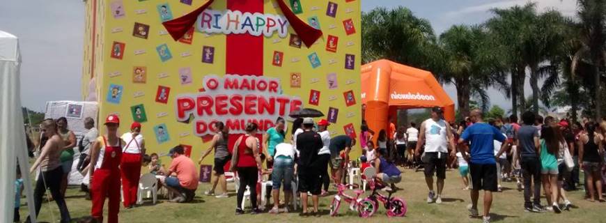 Foto pessoas caminhando em um parque com uma caixa gigante de presente da Ri Happy.