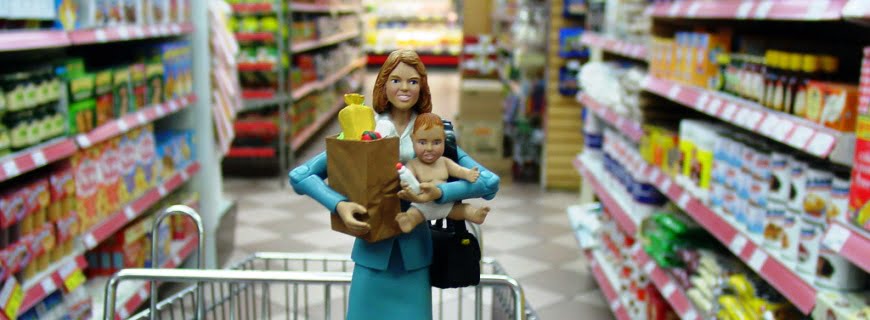 Foto de um boneco de uma mulher segurando em sua mão direita uma sacola de compras e com a sua mão esquerda segura um bebê, o boneco está em meio a prateleiras de supermercados.