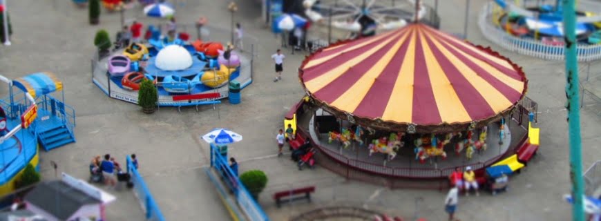 Foto de um parque de diversões sendo visto de cima, tem vários brinquedos diferentes, pessoas caminhão entre esses brinquedos.