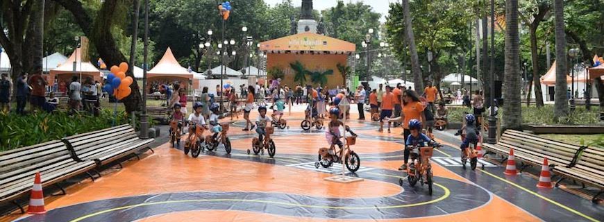 Foto de crianças sentadas em bicicletas, elas andam sobre uma pista feita em meio a uma praça.