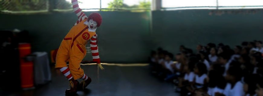Foto da mascote do McDonald's realizando uma peça em uma escola para crianças.
