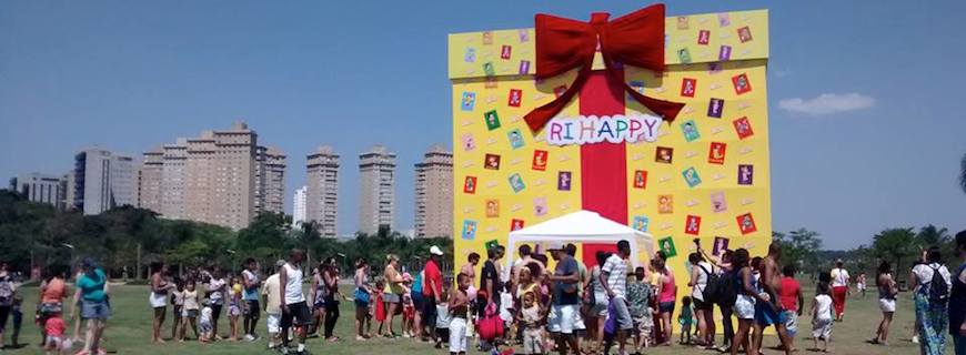 Foto de pessoas caminhando em uma praça com uma caixa de presente gigante da Ri Happy.
