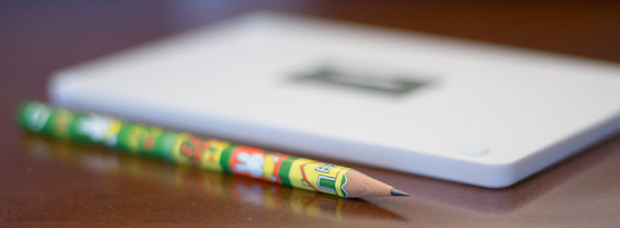 Foto de um lápis e um objeto semelhante a uma calculadora em cima de uma mesa de madeira.