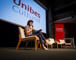 Foto de Ana Lucia Villela em um palco sentada em um cadeira falando ao microfone.