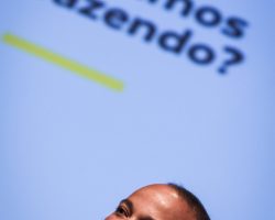 Foto de Pedrinho Fonseca segurando um microfone, ao fundo um telão está sendo projetado uma frase: O que estamos fazendo?