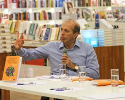 Calixto Salomão, sentado está falando em um microfone enquanto gesticula, em cima da mesa está o livro "Autorregulação da publicidade infantil no Brasil e no mundo".