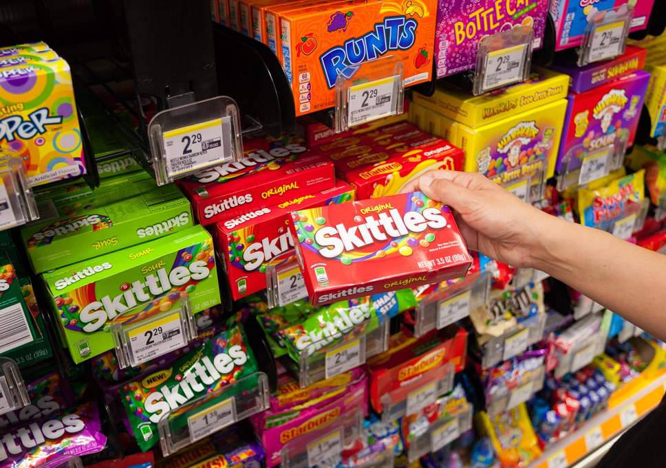 Foto com vários doces em uma prateleira de supermercado, uma pessoa está segurando uma caixa de um dos doces.