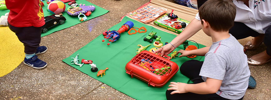 imagem com um menino sentado no chão, em frente alguns brinquedos em cima  de um papel verde, aparentemente sendo orientado por um adulto.