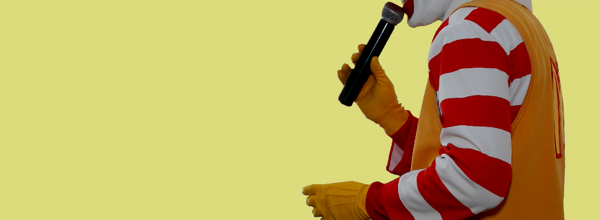 em fundo amarelo, foto em detalhe do palhaço Ronald McDonald segurando um microfone como imagem de apoio ao texto sobre show do ronald