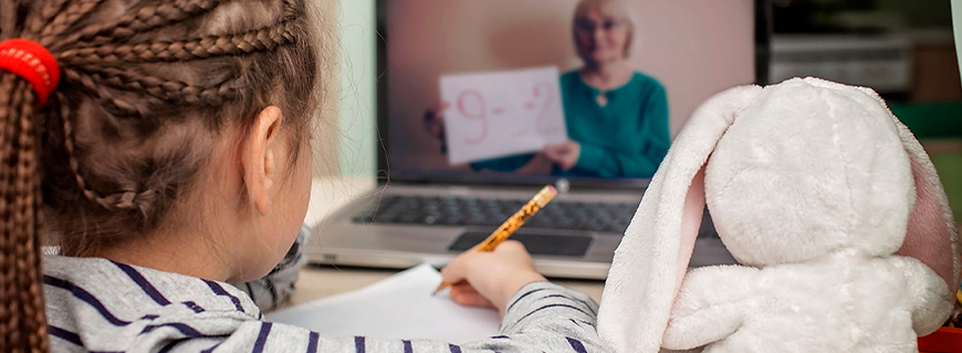 Foto de uma criança de costas em frente a um computador, segurando um lápis e um caderno e, ao lado direito dela, um bichinho de pelúcia também olhando o computador como imagem de apoio ao texto sobre a escola no mundo digital
