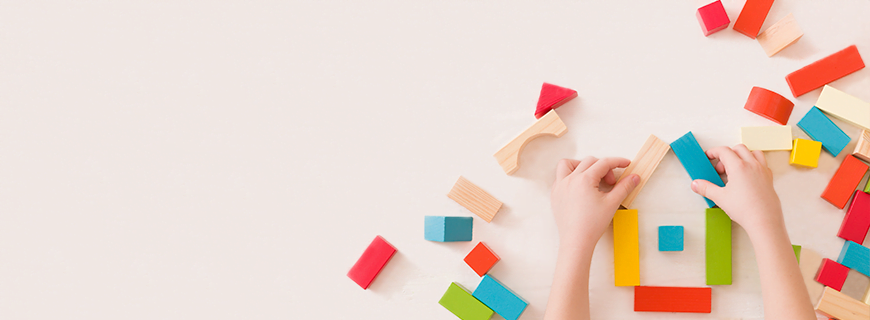 foto de mãos de uma criança brincando com blocos de montar de madeira coloridos em fundo bege claro como imagem de apoio ao texto sobre brincadeiras criativas