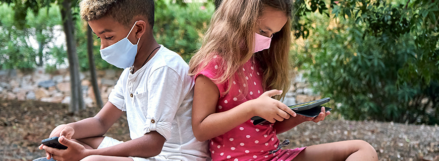 foto de duas crianças sentadas em um banco de um parque mexendo em um celular e um tablet
