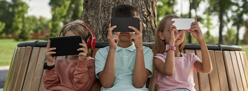 foto de três crianças sentadas em um banco em um parque usando celulares