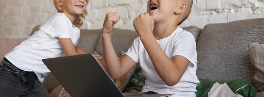 Menino branco sentado com um computador no colo e com os dois braços levantados em sinal de comemoração. Ao lado dele está uma menina branca sorrindo.