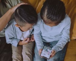 Na imagem, duas crianças usam um celular com um adulto responsável observando. A foto ilustra matéria sobre consulta pública sobre uso de telas realizada pelo Governo Federal.