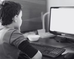 Imagem em preto e branco de um menino de costas olhando para o monitor de um computador. A imagem ilustra matéria sobre denúncia realizada no Habbo, jogo de simulação virtual.