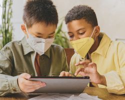Uma criança de etnia amarela e outra negra olham para um tablet. Elas utilizam máscaras. A imagem ilustra matéria sobre lançamento de cartilhas voltadas à defesa de crianças e adolescentes na Internet.